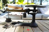 Side view of antique balance scale showing the crow's foot brass beam, circular cast iron weighing platform, and cast iron base with adjustable weights hung underneath.