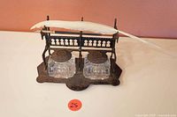 Front view of cast iron desk set holder with two clear glass inkwells with metal lids closed. White feather quill and black fountain pen resting on top.
