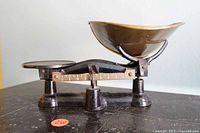 Side view of antique balance scale showing cast iron base and brass weighing pan, with graduated weight indicator visible.