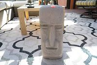 Front view of a gray concrete Easter Island Moai statue on a patterned outdoor rug with patio furniture in the background. The statue is roughly 7.5 x 6 x 18 inches with minimalist carved features and a red circle sticker on top.