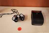 Front angled view of binoculars with neck strap and black storage case with red flap.