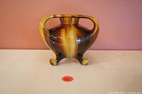 Front view of vintage pottery vase showing three legs, two handles and glossy brown and yellow glazing.