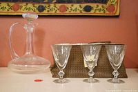 Clear glass swan decanter with stopper in background with 3 cut crystal glasses in front and woven zippered storage bag behind