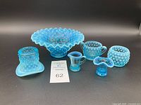 Photo of six vintage Fenton Blue Opalescent hobnail glass items arranged on a black background including bowl, cup, jar, creamer, candle holder and small vase.