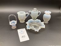 Five pieces of vintage Fenton glass displayed on a black background, including hobnail sugar bowl with lid, creamer, small basket with handle, bud vase, and candy dish.