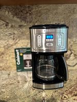 Photo showing the front view of the Cuisinart CBC-7000PCFR coffee maker on a kitchen countertop with digital display and control buttons, also showing the original box behind it.
