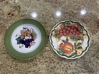 Image of the pair of vintage hand-painted ceramic plates placed on a granite countertop, showing detailed fruit motifs and green accents.