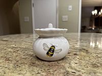 Front view of a white fine bone china honey pot with a bee illustration on the body and a scalloped edged lid with a knob handle.