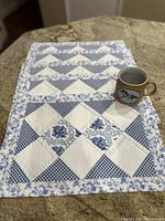 Set of blue and white floral quilted placemats stacked with a ceramic horse design mug on top.