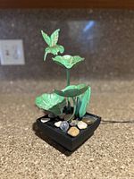 Angled view of vintage style metal sculpture featuring green patina hummingbird and lotus flowers on black square base with stones.