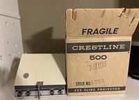 Side view of the Crestline 500 Deluxe slide projector next to its original cardboard box labeled 'Fragile' and 'Crestline 500 Deluxe' with stock number 6258.