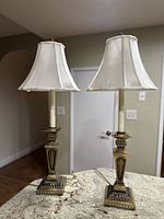 Pair of vintage brass candlestick table lamps with cream fabric shades shown on a marble surface and hardwood floor background.
