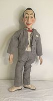 Full standing view of Pee-wee Herman doll in gray suit, white shirt, red bow tie, beige shoes, with painted face and styled hair.