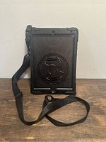 Front view of black protective iPad case with shoulder strap attached, showing rugged design and hand strap hub.
