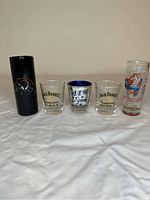 Front view of five shot glasses arranged in a row, showing various designs including Jack Daniel's Tennessee Honey branding, floral art, and a black glass.