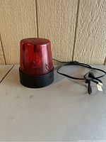 Red rotating beacon light with black base and cord placed on table against wood panel wall.