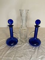 Front view showing the clear glass vase centered with the two blue glass bottles on either side, all on a white cloth background.