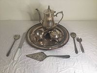 Silver-plated tea set on round WM Rogers tray including teapot, cream pitcher, sugar bowl, and five utensils alongside an additional oval WM Rogers tray.