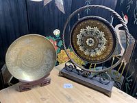 Photo showing the 14-inch brass bowl with dragon engravings on a wooden stand next to the 19-inch decorative brass disc on a black iron frame stand
