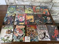 Photo showing all 15 comic books laid out in three rows on a wooden surface in front of a stone wall. Covers are visible, mostly superhero and fantasy genre comics including Madame Xanadu, Cinder & Ashe, Max Mage, Wild Cards, Darkhawk, Score, Big Hero, The Great Unknown.