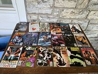 Overview of 18 vintage comic books arranged in rows on a wooden surface outside a stone house wall.