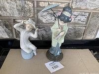 Both vintage Lladro porcelain figurines positioned side-by-side on a black piano surface with stone wall background.