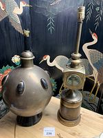 Front view of the two lamps on a wooden surface with a decorative black background featuring crane illustrations. The spherical brass lamp is on the left, with black base and oval protrusions. The railroad lantern style lamp is on the right with clear square glass panes and a tall chimney.