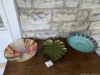Three large decorative fruit bowls displayed: green leaf-shaped silver plate, multicolored carnival glass, and blue scalloped glass bowl.