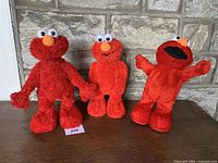 Front view of three vintage Tickle-Me-Elmo plush dolls showing red fur, orange noses, and white eyes on a wooden surface with stone wall background.