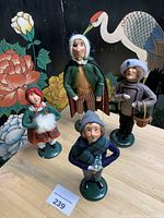 Front view of four Byers Choice Carolers Nutcracker figurines dressed in winter clothing with painted faces and fabric accessories placed on table with decorative floral and bird backdrop.