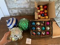 Photo displaying a large box containing about 40 small Christmas ball ornaments in red, gold, green, and blue, a smaller box with approximately 12 assorted color ornaments, and 3 large decorative balls in varying colors and intricate patterns.