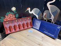 Six Cristal D'Arques crystal wine glasses arranged in original coral cardboard tray, with the blue branded original box beside them on wooden surface.