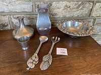 Six-piece vintage pewter set on wooden surface against stone wall, includes bowl, utensils, cup, ladle, and decorative item.