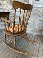 Side angled view showing wooden rocking chair with spindled backrest and floral seat cushion, placed on stone floor and in front of stone wall.