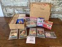 View of entire lot showing box of assorted vintage baseball trading cards, several displayed cards in plastic protectors, plastic bag of loose cards, and unopened 1986 football and 1991 baseball trading card packs.
