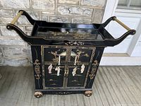 Full view of black lacquered wheeled tea cart cabinet with painted Asian-style figures and floral designs on cabinet and sides.