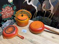 Three-piece set of vintage enameled cast iron cookware, volcanic orange color, displayed on wooden surface with decorative screen backdrop.