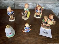 All six Goebel Hummel figurines and porcelain bell arranged on table showing variety and detail