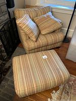 Full view of large armchair with matching ottoman and two pillows, positioned next to fireplace and window in room corner.