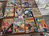 Image of all 12 comic issues laid out on wooden table showing front covers, including Isis, Wonder Woman, Batman, G.I. Joe, Thor, World of Krypton, Super Villains and others.