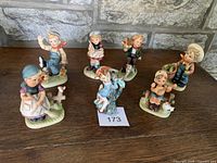 Set of seven vintage Napco Ware figurines displayed on wood surface against stone wall background. Poses include children with animals and objects such as baskets and garden tools.