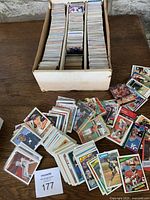 Photo showing a cardboard box filled with thousands of sports trading cards, some spilled out on a wooden table surrounding the box.