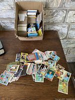 Photo showing a cardboard box filled with mixed loose baseball trading cards along with a spread of cards on a wooden surface.