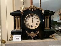 Frontal image of the WM.L Gilbert antique mantel desk clock showing its two brass pillar legs, black body, and ornate gold decorations