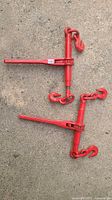 Two red ratchet chain binders laid out on the ground, showing full length from hooks to ratchet handles.