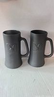 Front view of two black matte glass mugs standing side by side, showing handles and Playboy bunny logos.