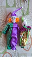 Front view of two clown porcelain dolls, one in green and purple satin with orange hair, the other in gold and green satin with white hair.