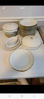 Stacked dining plates, covered soup containers, narrow plate, and a gravy dish on table, showing white porcelain with gold rim trimming.