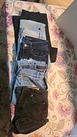 Five pairs of pants and shorts arranged on a patterned bedspread. Several are denim, varying shades from light blue to black. The items include pants and shorts, sizes noted as 25.