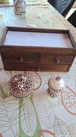 Wooden jewelry box with three drawers and two decorative ceramic trinket boxes sitting on a patterned tablecloth.